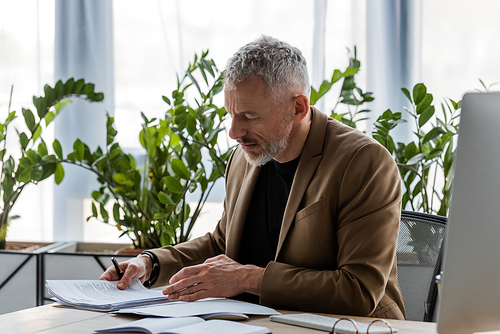 bearded businessman looking at document while sitting at desk in office