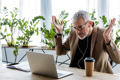 confused businessman in glasses and earphones showing shrug gesture and looking at laptop while having video call