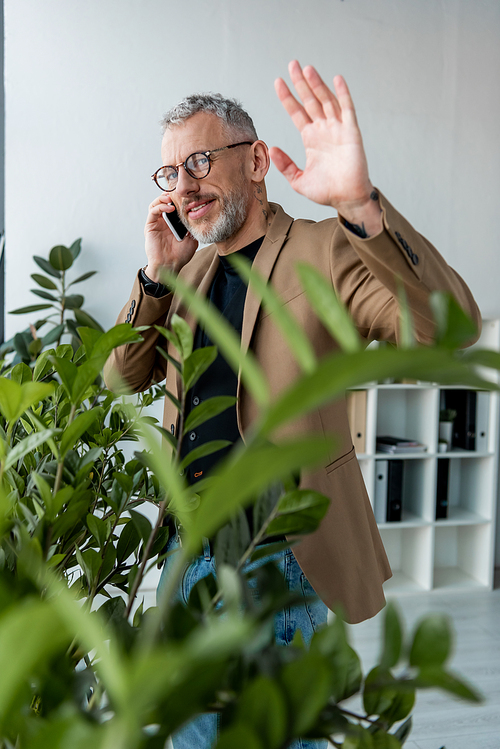 selective focus of happy businessman waving hand while talking on smartphone near plants