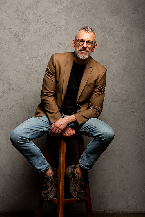 bearded businessman in glasses  and sitting on stool on grey