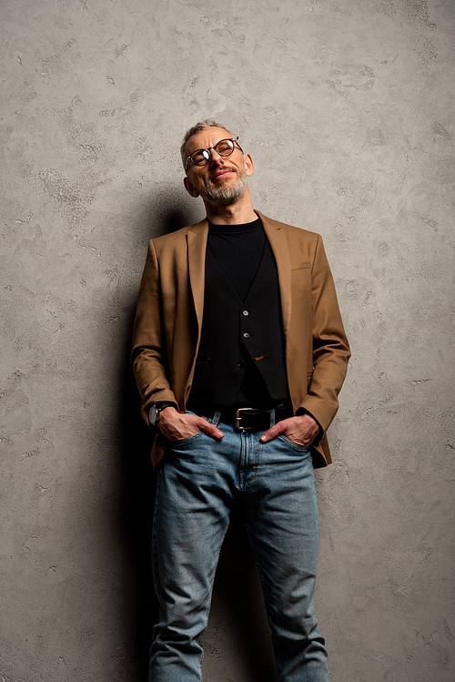 handsome businessman in glasses standing with hands in pockets on grey