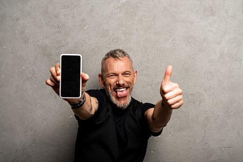 selective focus of happy man sticking out tongue and holding smartphone with blank screen while showing thumb up on grey
