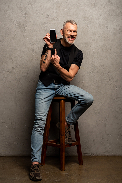 happy man with tattoo pointing with finger at smartphone with blank screen on grey