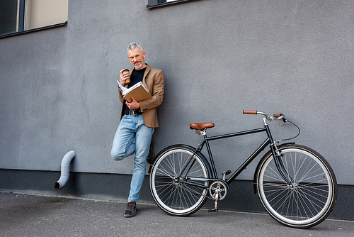 handsome businessman in blazer and jeans standing near bicycle while holding paper cup and folder