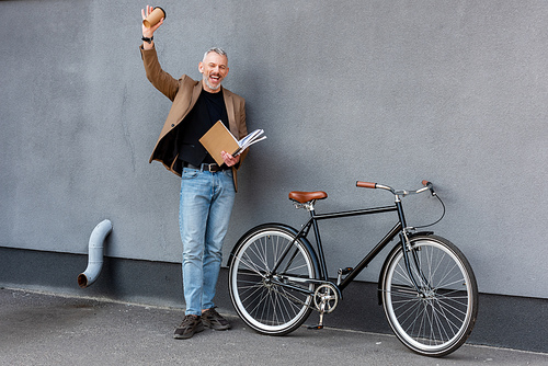happy businessman waving hand while holding paper cup and folder and standing near bicycle