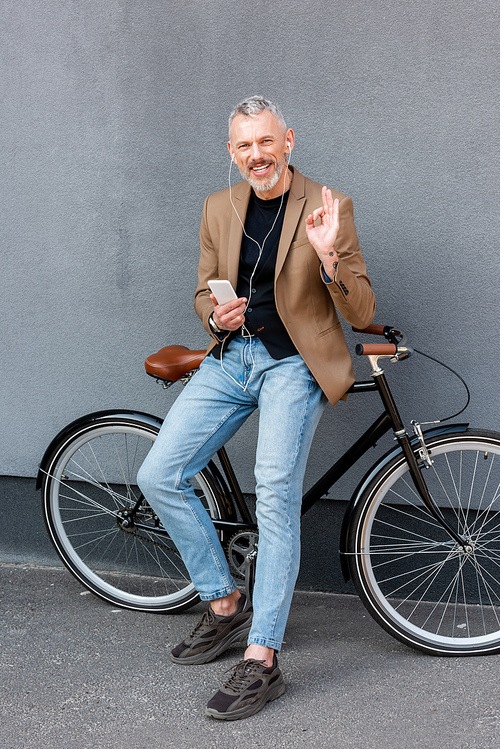cheerful businessman in earphones listening music while showing ok sign and holding smartphone near bicycle outside