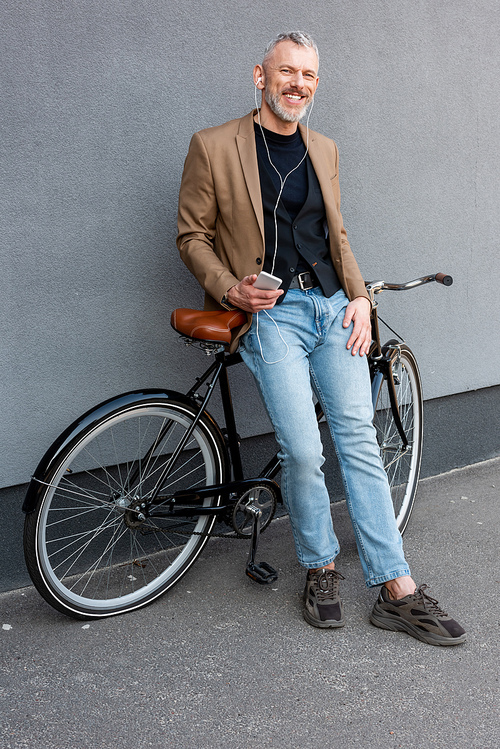 cheerful businessman in earphones listening music while holding smartphone and standing near bicycle outside