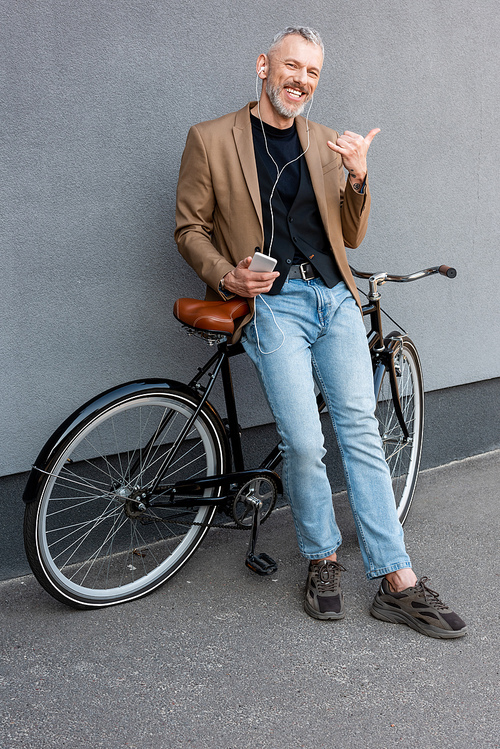 cheerful businessman in earphones listening music while showing call me gesture and holding smartphone near bicycle outside