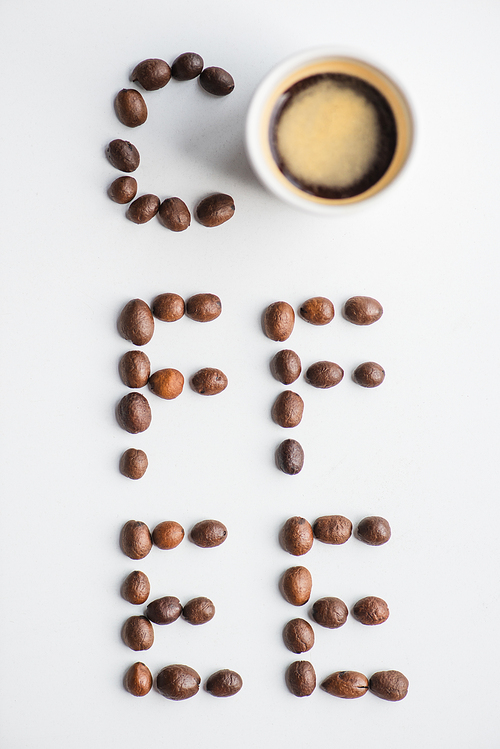 top view of coffee lettering made of beans near cup on white surface