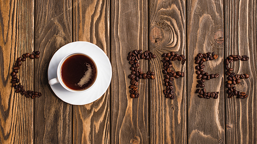 top view of cup of coffee on saucer and coffee lettering made of beans on wooden surface