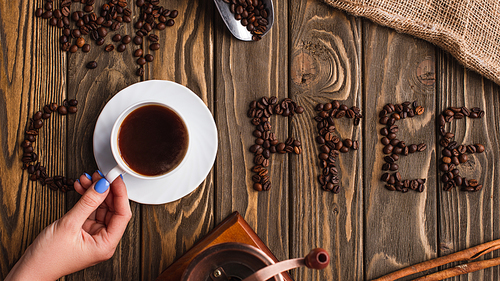 cropped view of woman holding cup of coffee on saucer near coffee lettering made of beans on wooden surface