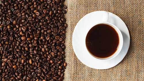 top view of cup of coffee on saucer with coffee beans and sackcloth on background