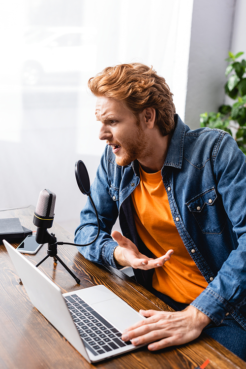 tense broadcaster in denim shirt gesturing while talking in microphone near laptop