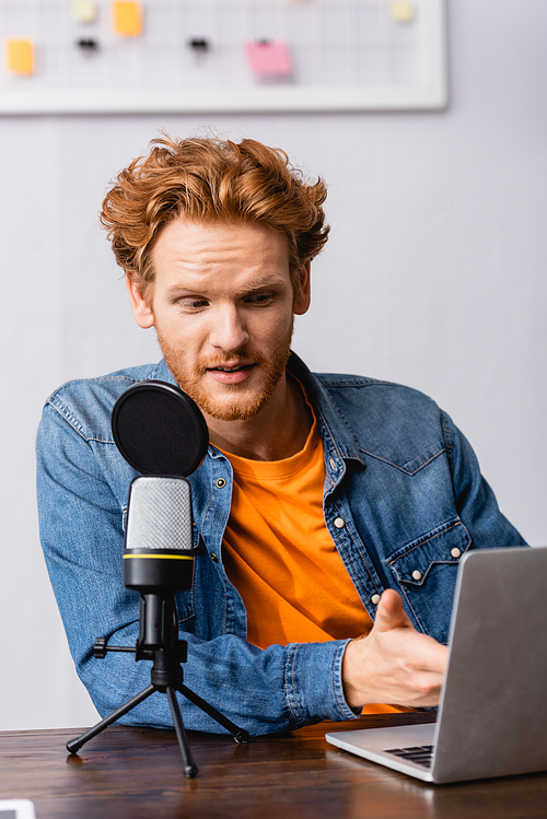 young broadcaster in denim shirt pointing with hand at laptop while speaking in microphone