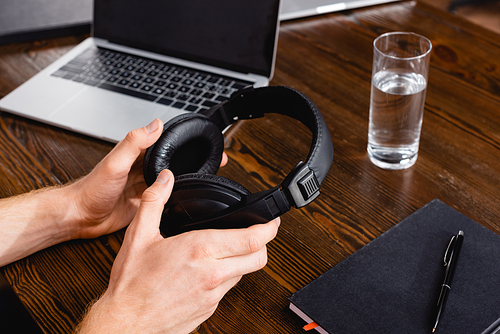 partial view of freelancer holding wireless headphones near laptop, glass of water and notebook with pen