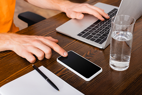 partial view of freelancer using laptop and touching smartphone with blank screen near glass of water and notebook