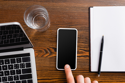 cropped view of freelancer touching smartphone with blank screen near laptop, notebook and glass of water