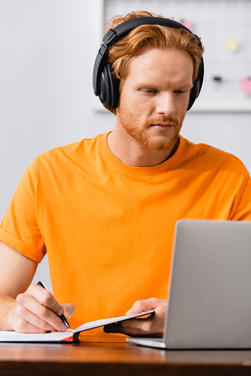 concentrated freelancer in wireless headphones writing in notebook while looking at laptop