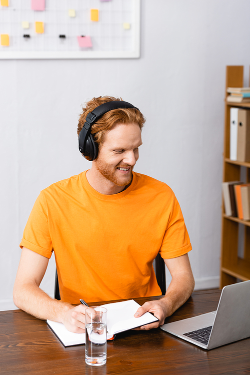 young freelancer in wireless headphones writing in notebook while looking at laptop