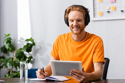 redhead student in wireless headphones using digital table while writing in notebook at home