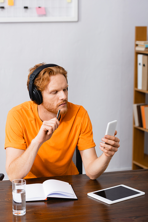 pensive student in wireless headphones holding pen while using smartphone near digital tablet with blank screen