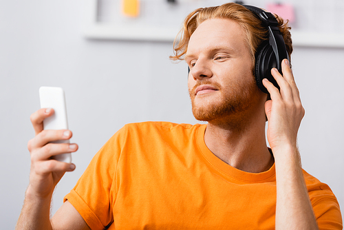 selective focus of dreamy man touching wireless headphones while holding smartphone