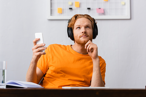 dreamy redhead man in wireless headphones looking up while holding smartphone