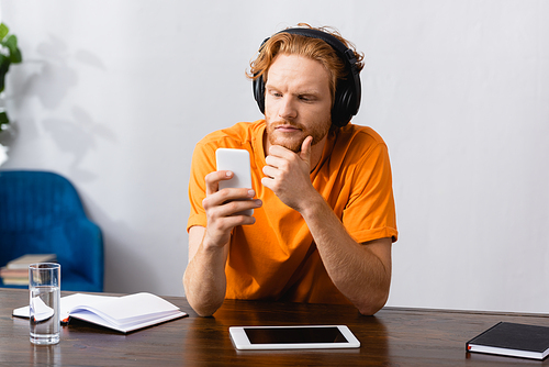 thoughtful student in wireless headphones touching chin while using smartphone at home