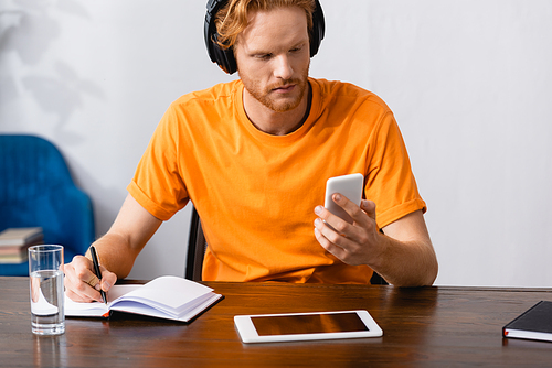 concentrated student in wireless headphones using smartphone and writing in notebook