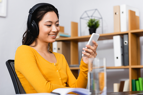 selective focus of brunette asian woman listening podcast in wireless headphones while using smartphone at home