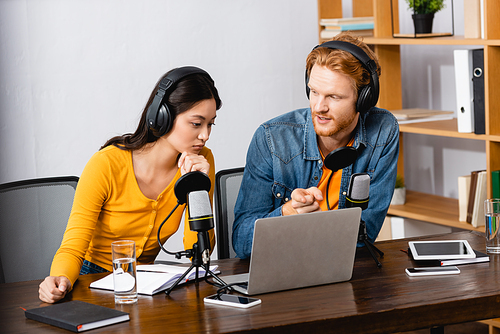 radio host pointing with finger at laptop near thoughtful asian colleague in wireless headphones