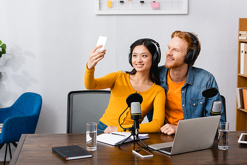 asian broadcaster in wireless headphones taking selfie on smartphone with young colleague