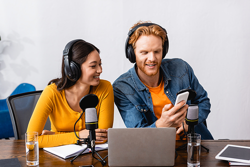 young broadcaster in wireless headphones using smartphone near asian colleague