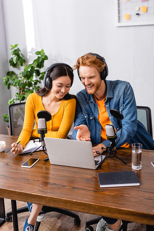 young broadcaster pointing with hand at laptop near asian colleague in wireless headphones