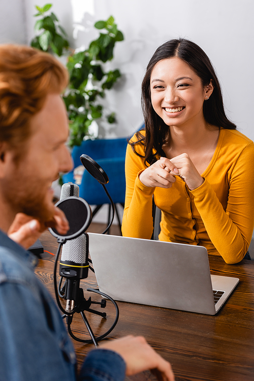 selective focus of excited asian interviewer looking at guest in radio studio