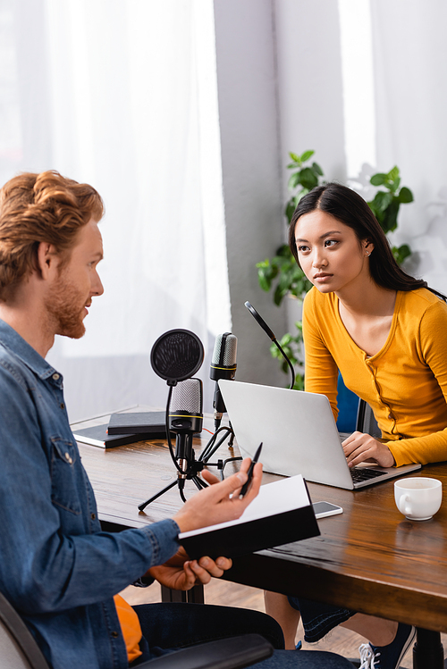 redhead man holding notebook and pen while talking to young asian interviewer in studio