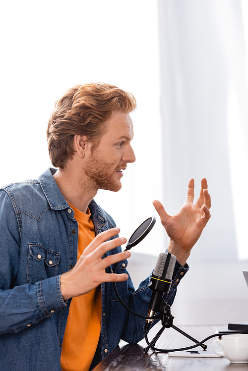 redhead, bearded broadcaster gesturing while talking near microphone at workplace