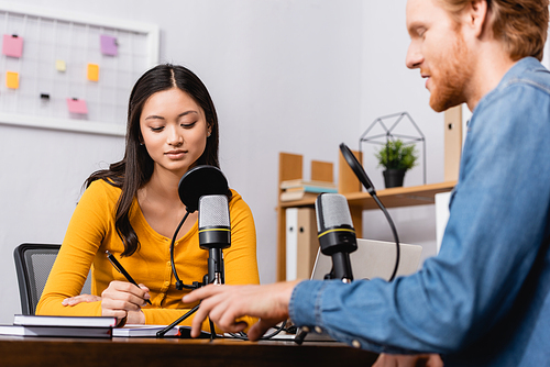 selective focus of young asian broadcaster holding pen during interview with redhead man