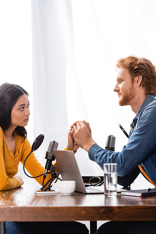 young redhead radio host holding hands with sad asian woman during interview in studio