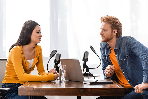 redhead radio host interviewing brunette asian woman near microphones and laptop in studio