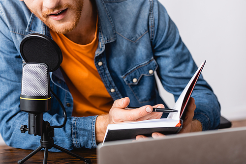 partial view of bearded broadcaster in denim shirt holding notebook and pen while speaking in microphone