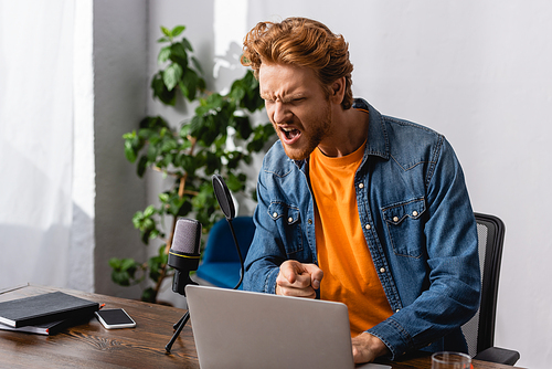 angry broadcaster in denim shirt pointing with finger while screaming in microphone near laptop