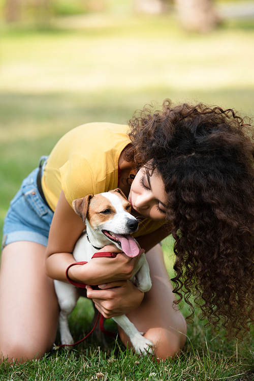 Selective focus of young woman sitting on grass and kissing dog