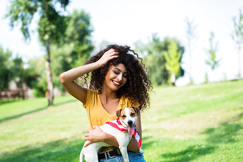 joyful woman touching curly hair while holding jack russell terrier dog in park