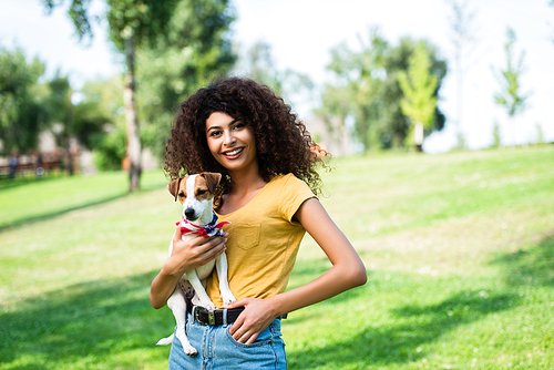 young woman in summer outfit standing with hand in pocket while holding jack russell terrier dog in park