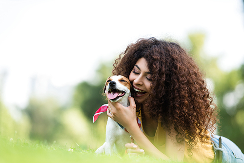 selective focus of excited, curly woman hugging jack russell terrier dog while lying on grass