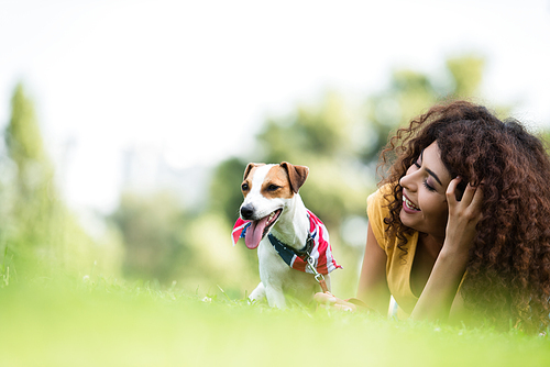 selective focus of curly woman laughing while resting with jack russell terrier dog on lawn