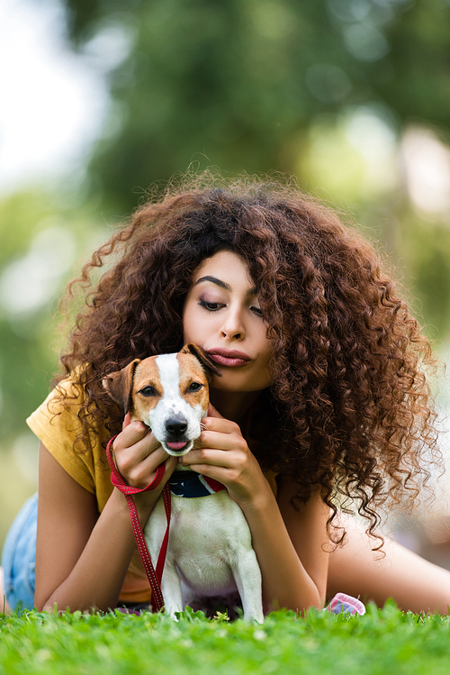 brunette, curly woman pouting lips while relaxing with jack russell terrier dog on green grass