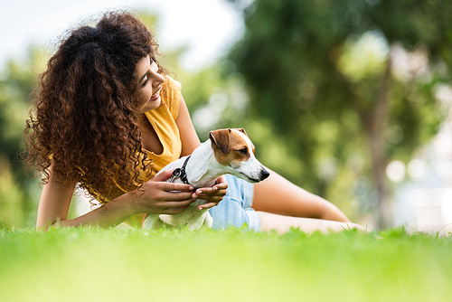 selective focus of curly woman looking away and laughing while lying on lawn with jack russell terrier dog