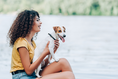 side view of excited woman in summer outfit sitting near lake with jack russell terrier dog